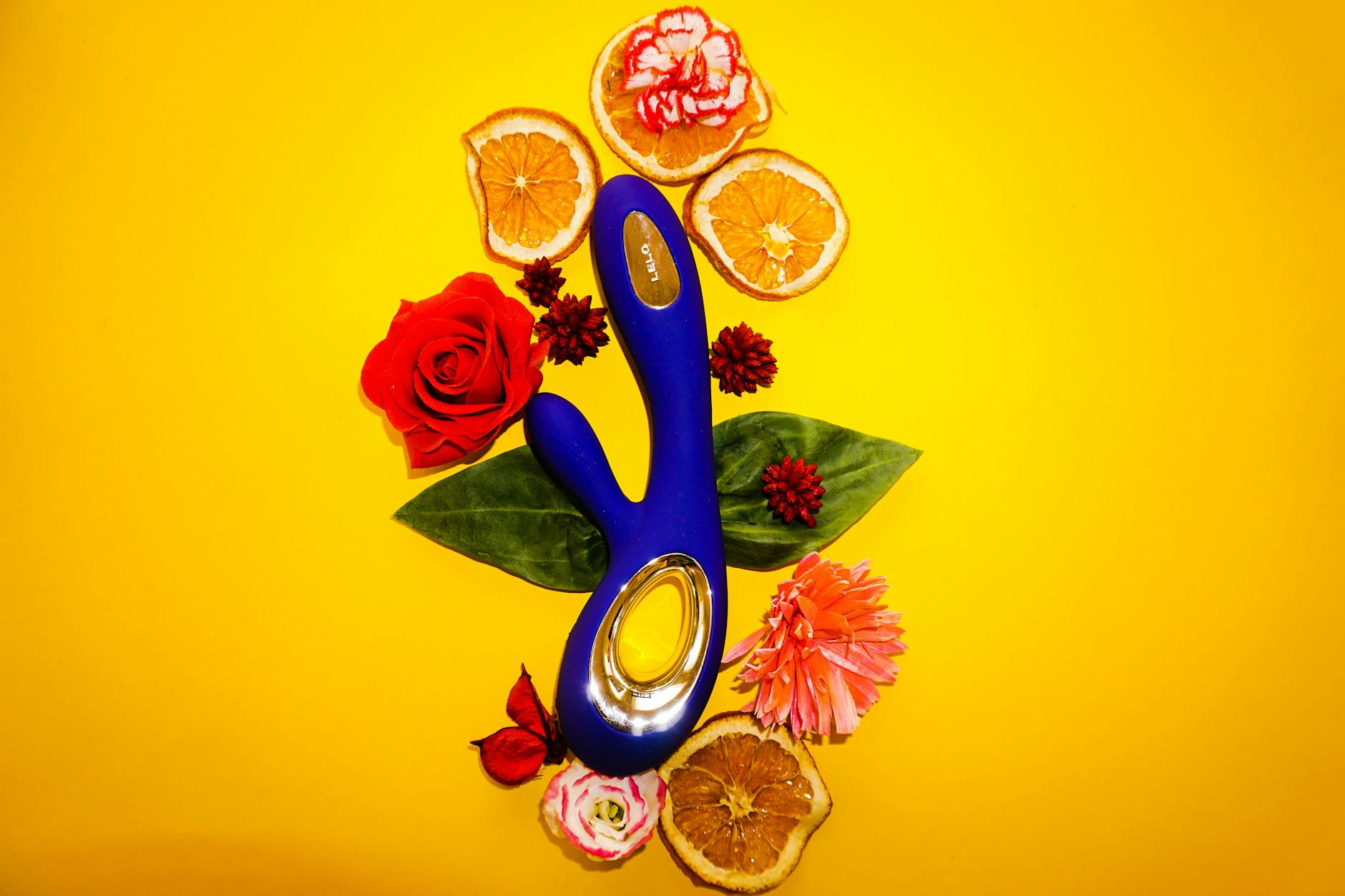 Blue vibrator surrounded by flowers and citrus on a bright yellow background, symbolizing mindful pleasure and sensory focus.