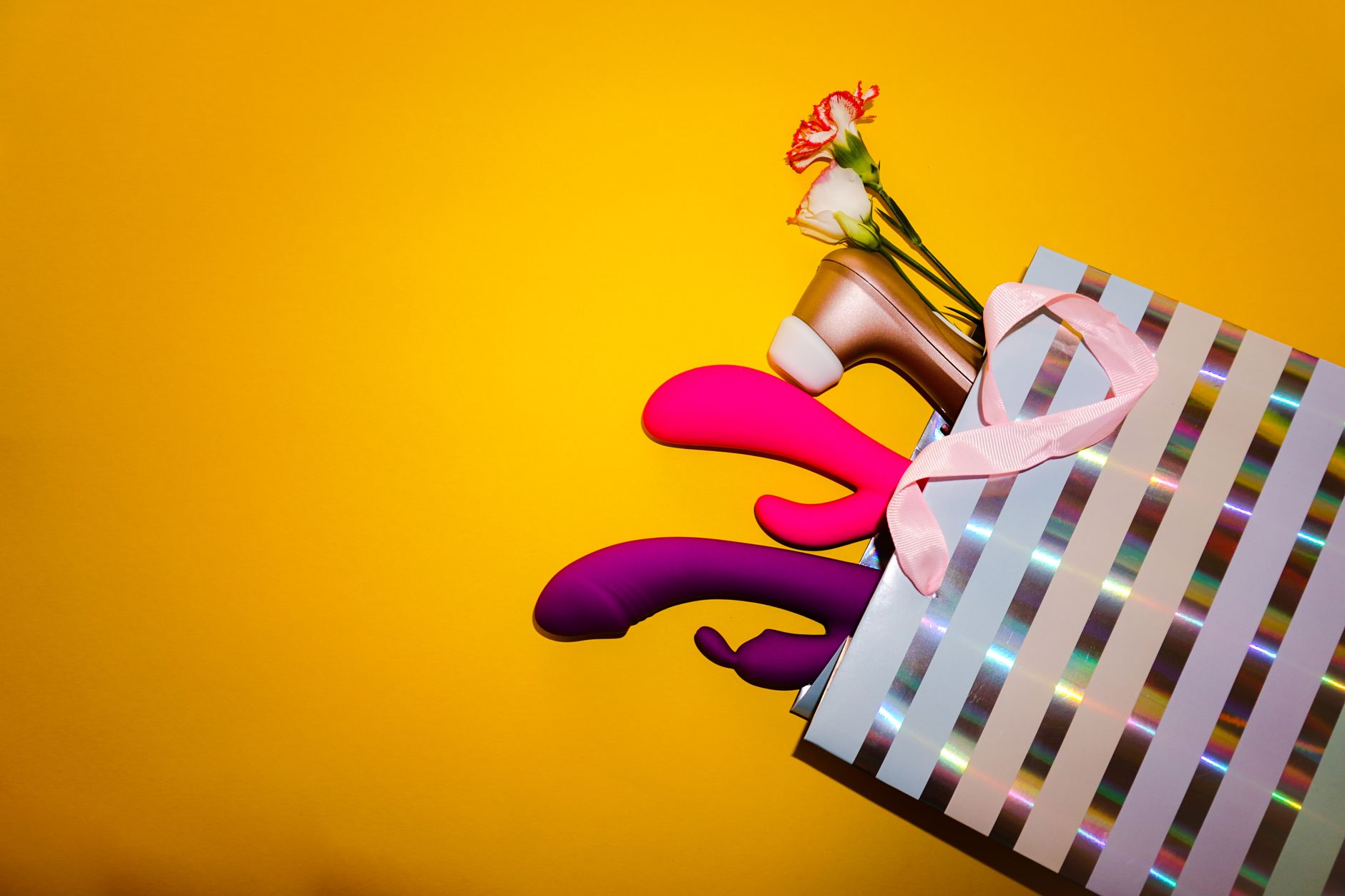 Colorful vibrators with flowers in a holographic gift bag on a bold yellow background